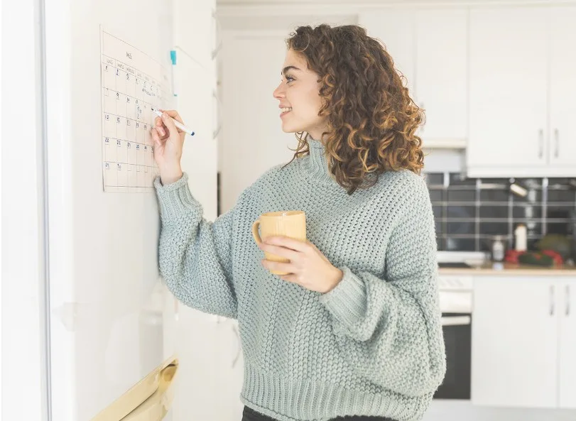 Young woman writing in a calendar on a whiteboard while holding a cup of coffee