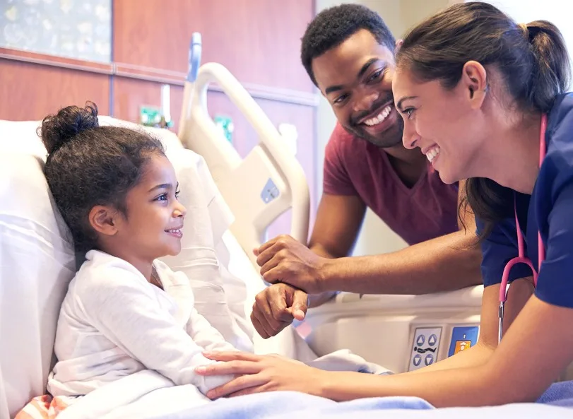 Man and a nurse smiling looking at their a child in a hospital bed while she looks back with a smile